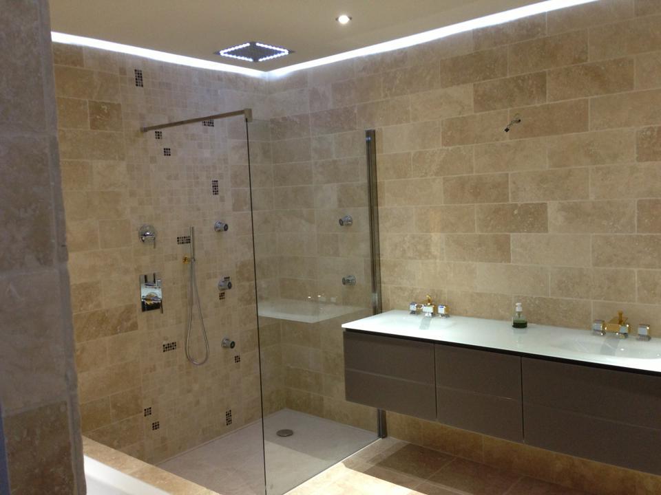 Salle de bains en travertin : douche à l'italienne vitrée, double vasque suspendue et corniche lumineuse LED